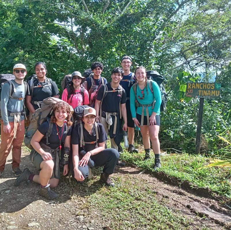 A group of nine young adults are posing for a photo on a trail. They are all wearing backpacks, suggesting they are hiking. The background features lush green vegetation and a sign that reads "Ranchos Tinamu". The group appears to be in good spirits, likely enjoying their outdoor adventure.